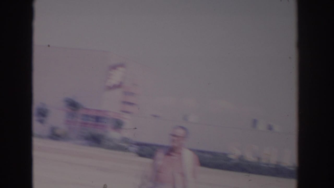 Man in glasses walking near parked cars and a large building with a blurred sign.