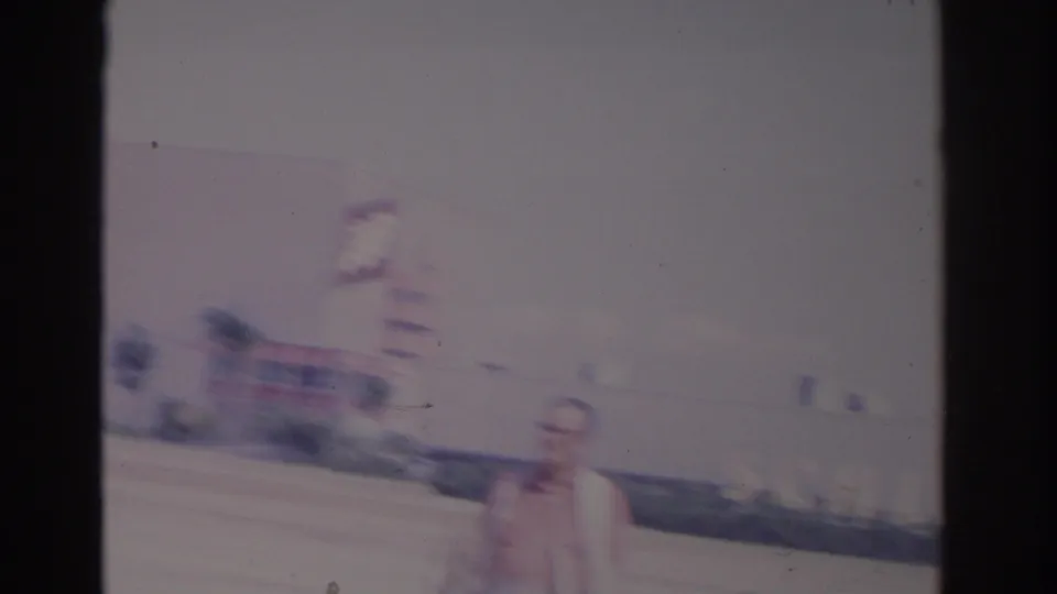 1962 Florida home-movie frame with an adult man in glasses walking past parked cars near a commercial building and visible signage.