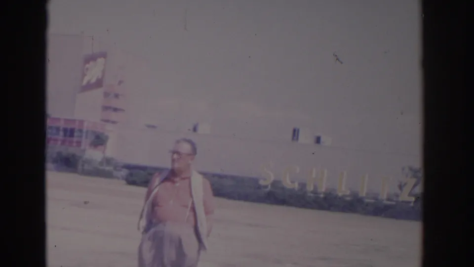 1962 Florida 8mm frame showing an adult man in glasses walking before a building with visible Schlitz signage and palm landscaping.