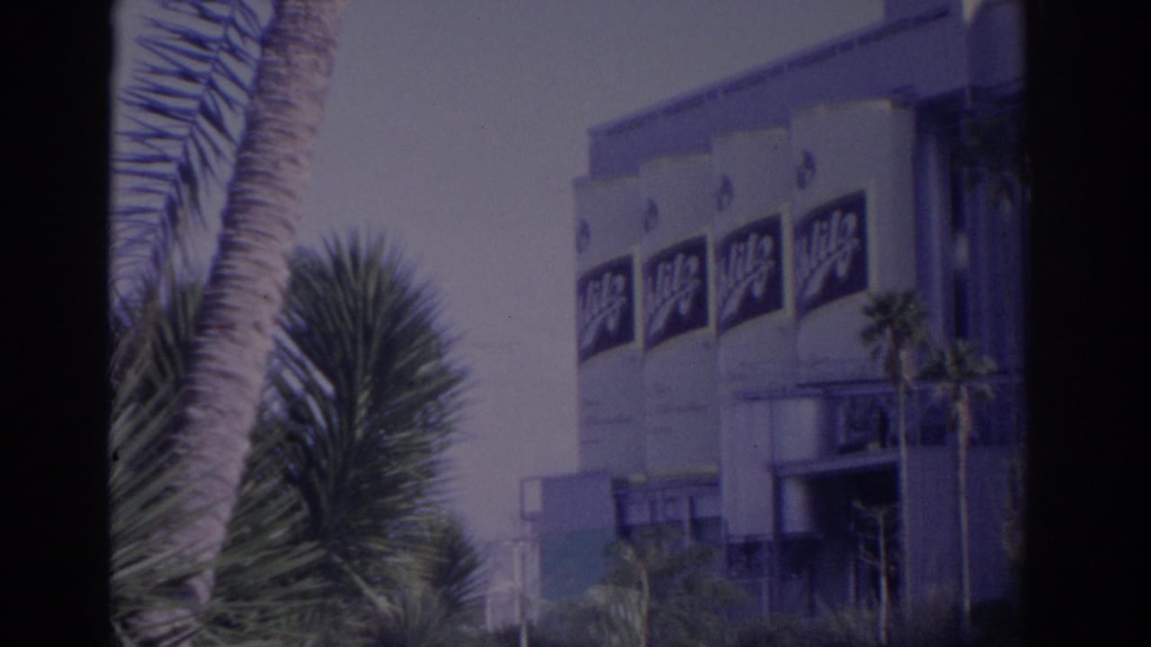 Branded industrial building facade partly framed by palms and dark film edges.