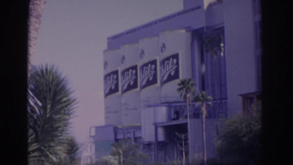 1962 Florida home-movie frame of a large branded building wall with repeated signs and tropical landscaping.