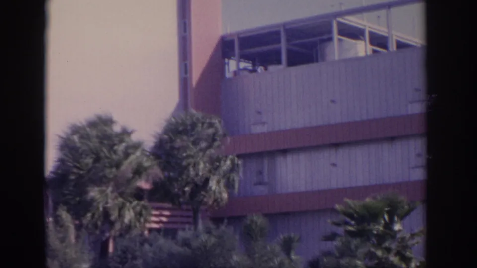 1962 Florida frame showing the building side with upper windows, horizontal red bands, and dense palm landscaping.