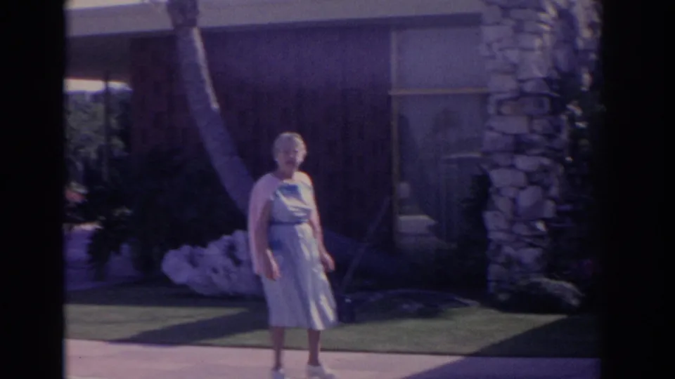 1962 Florida home-movie frame of an older woman walking past a lawn, palm trunk, stone pillar, and shaded building exterior.