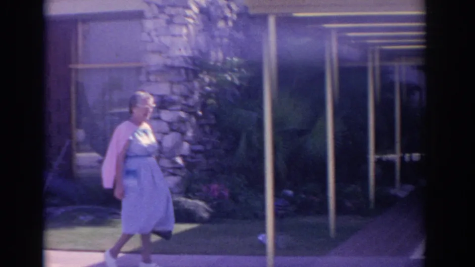 1962 Florida frame showing an older woman in a light dress and pink cardigan near glass doors, a stone pillar, lawn, and palms.