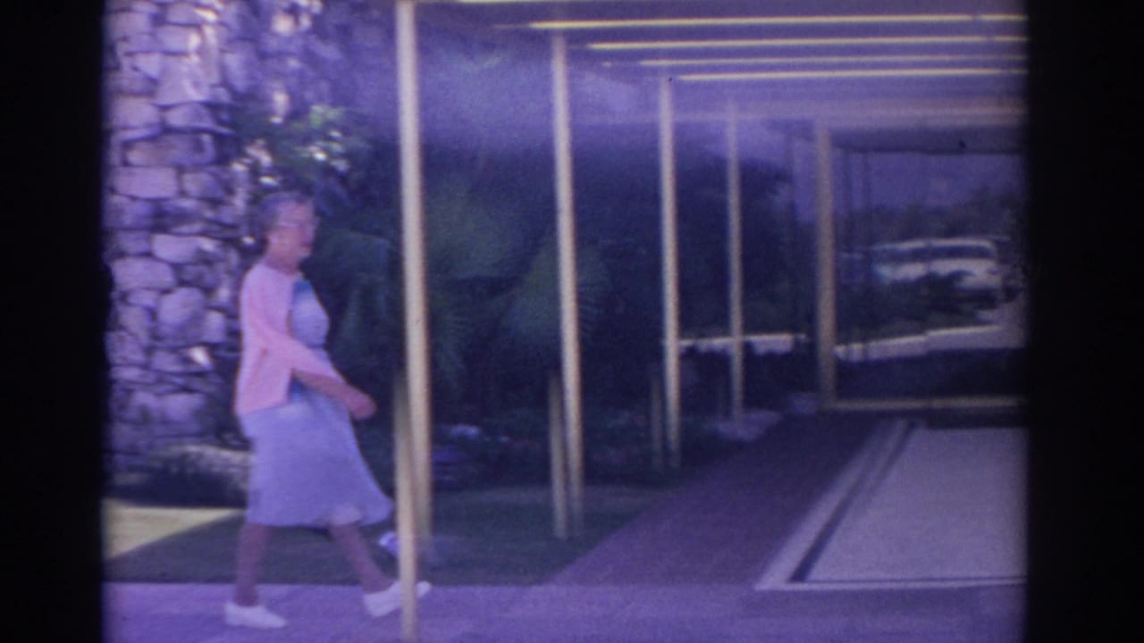 Woman in a blue dress and pink cardigan walks beside a stone pillar near glass doors.