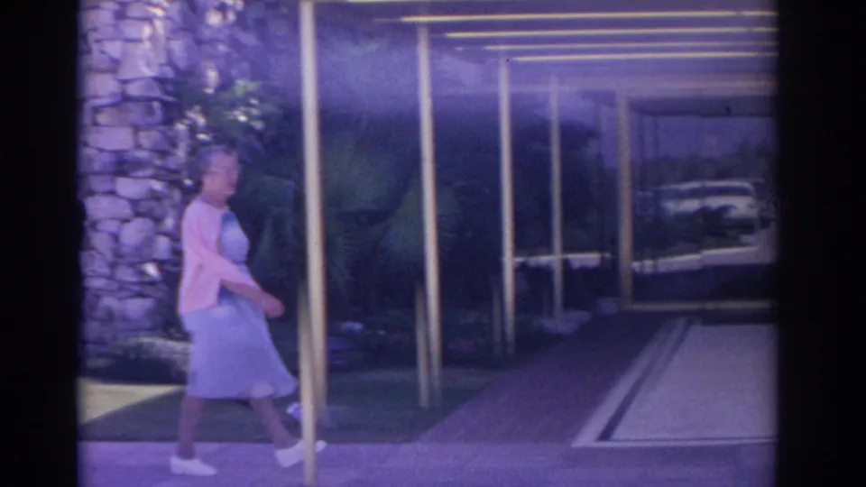 In Florida 1962 home-movie footage, a woman in a blue dress and pink cardigan walks along a sunny modernist entrance.