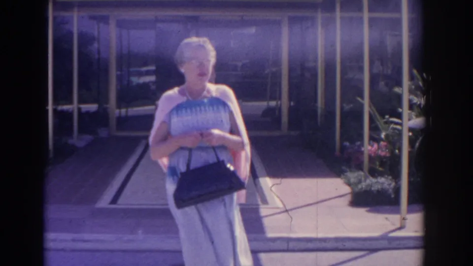 A woman in a blue patterned dress and pink cardigan approaches the camera, holding a black handbag outside a Florida entryway in 1962.