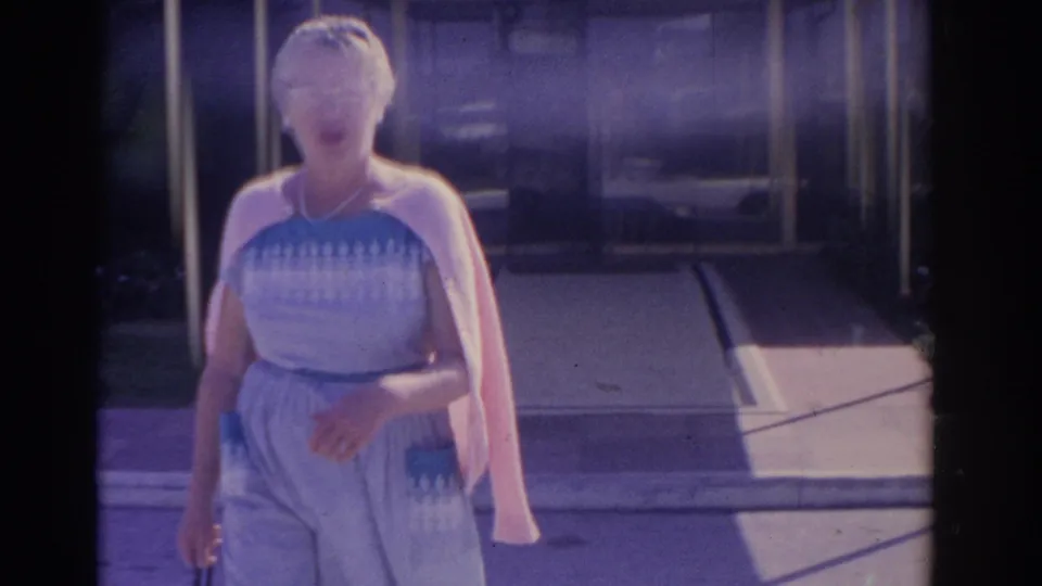 Closer 1962 Florida frame of the woman in a blue dress and pink cardigan walking toward the camera in strong sunlight.