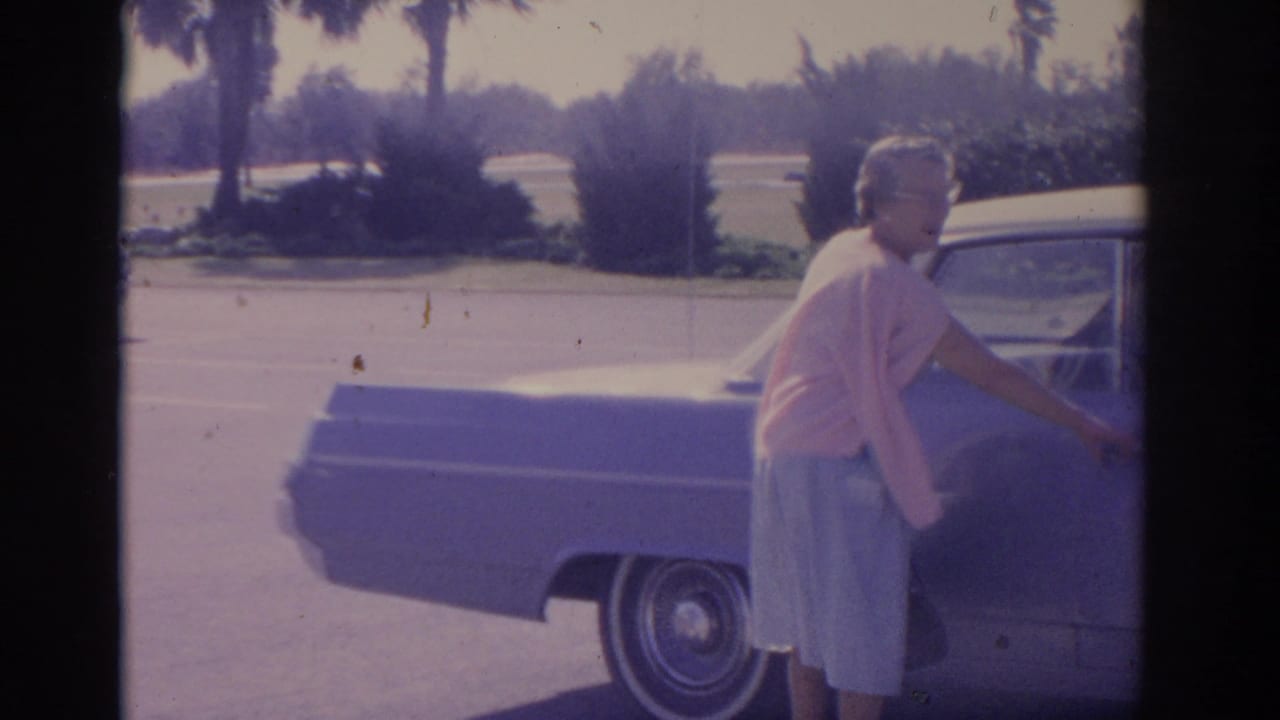 Woman in a pink cardigan opens the door of a large parked sedan in bright sun.