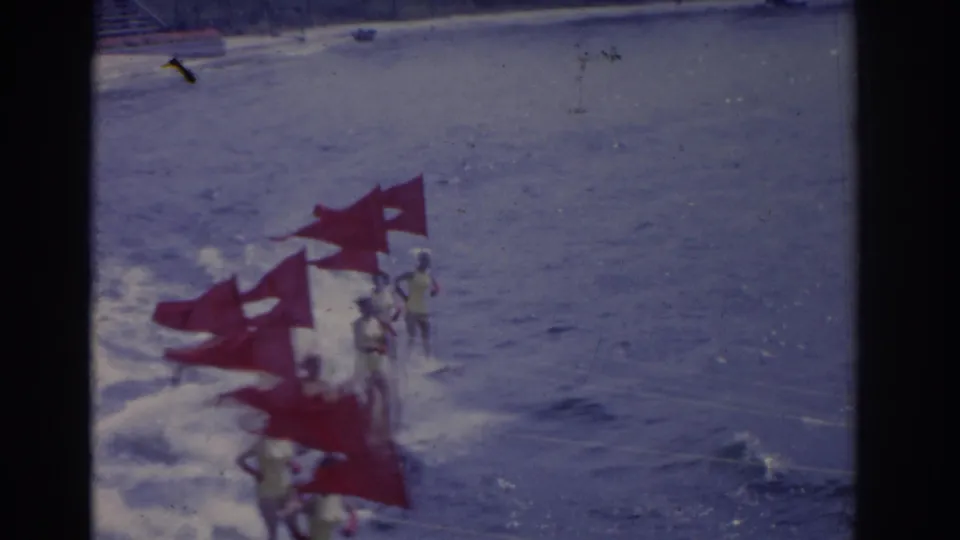 Surfers move through choppy surf near red triangular flags, with the water and shoreline visible in faded 8mm color.
