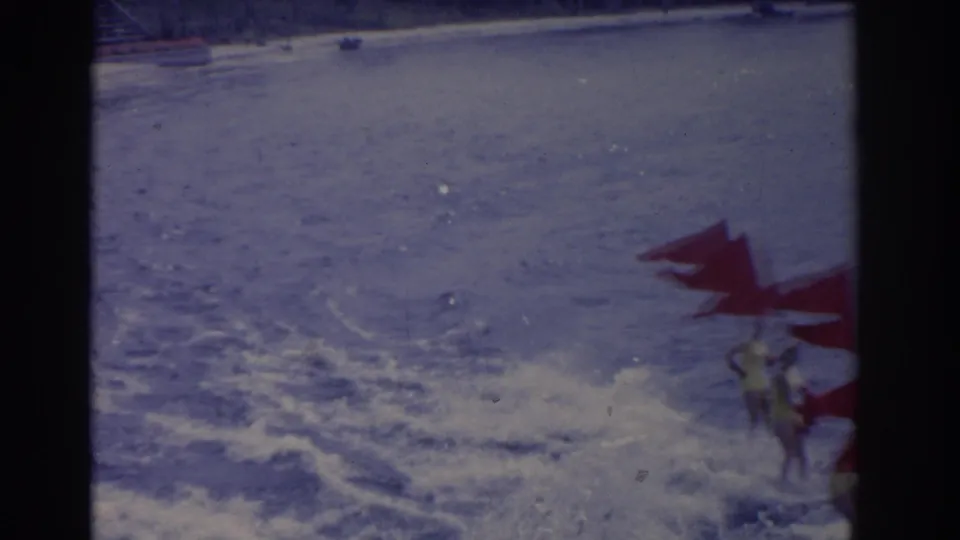 A handheld beach view shows red flags and partially visible surfers at the edge of foamy surf in archival 8mm footage.
