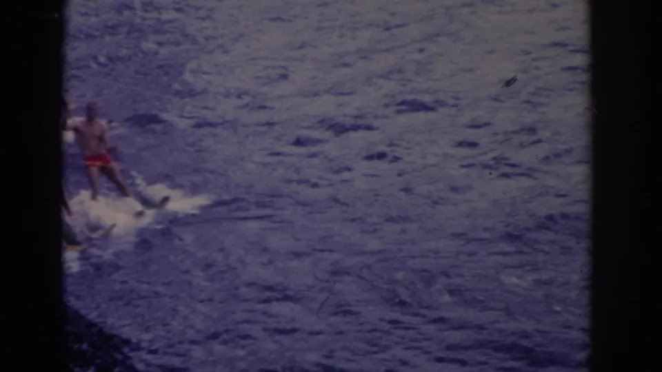 1962 Florida home-movie frame showing a distant man in red trunks water skiing over choppy blue water.