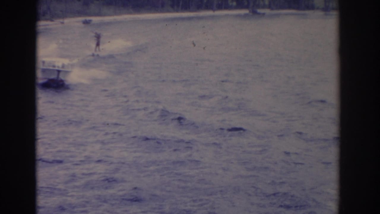 Water skier trails behind a small speedboat on a lake, framed by dark film borders.