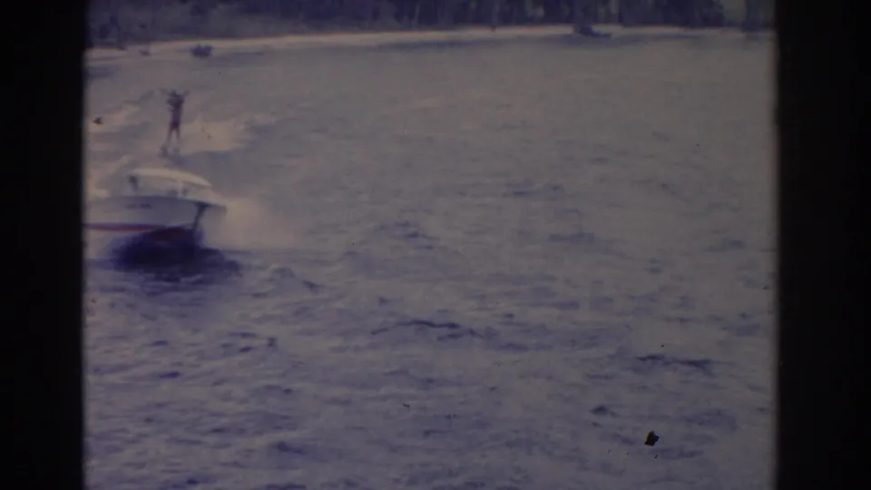 1962 Florida home-movie frame with a white speedboat and a distant water skier crossing the lake behind it.