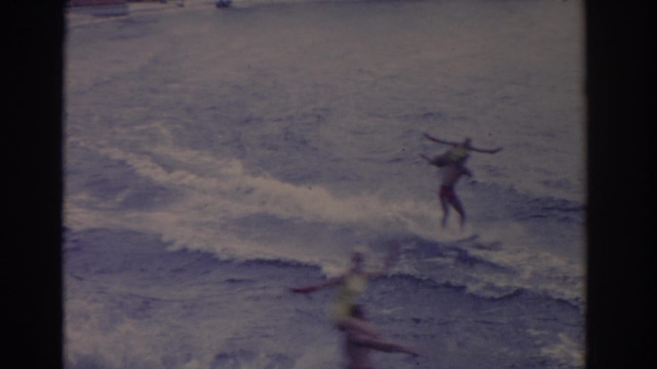 Water skier rides near a wake as a blurred foreground figure passes below.