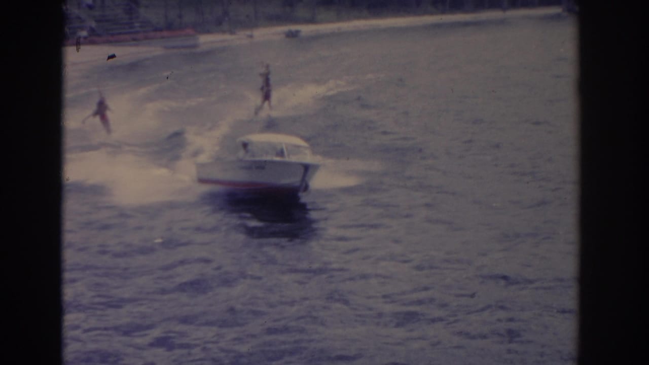 White motorboat towing water skiers across open water with a beach in the background.