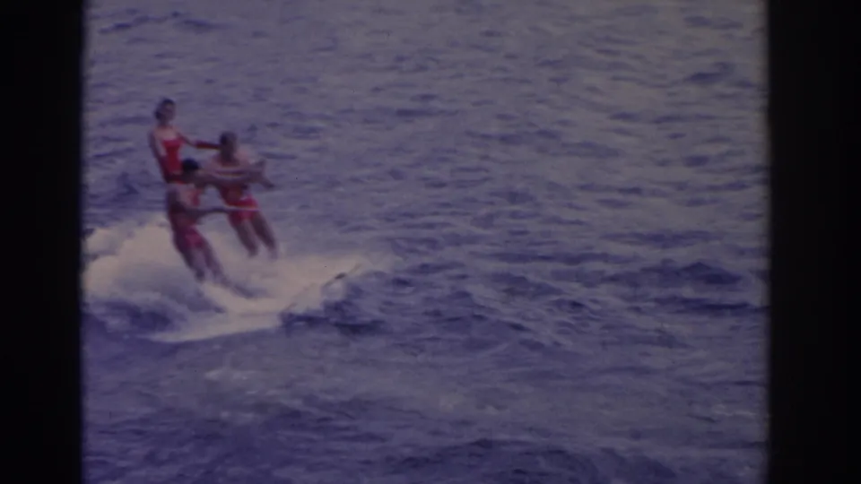Three people in red swimwear waterski together in 1962 Florida footage, leaning over choppy water with white spray below.