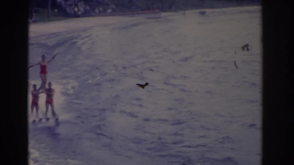 In 1962 Florida home-movie footage, three water skiers in red swimwear form a shoulder-balance stunt on open water.