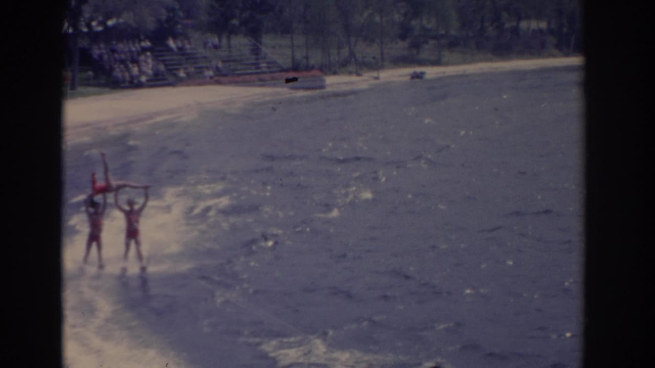 1962 Florida home-movie frame showing red-suited waterski performers forming a lift near shore, with beach seating behind them.