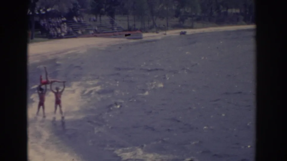 1962 Florida home-movie frame showing red-suited waterski performers forming a lift near shore, with beach seating behind them.