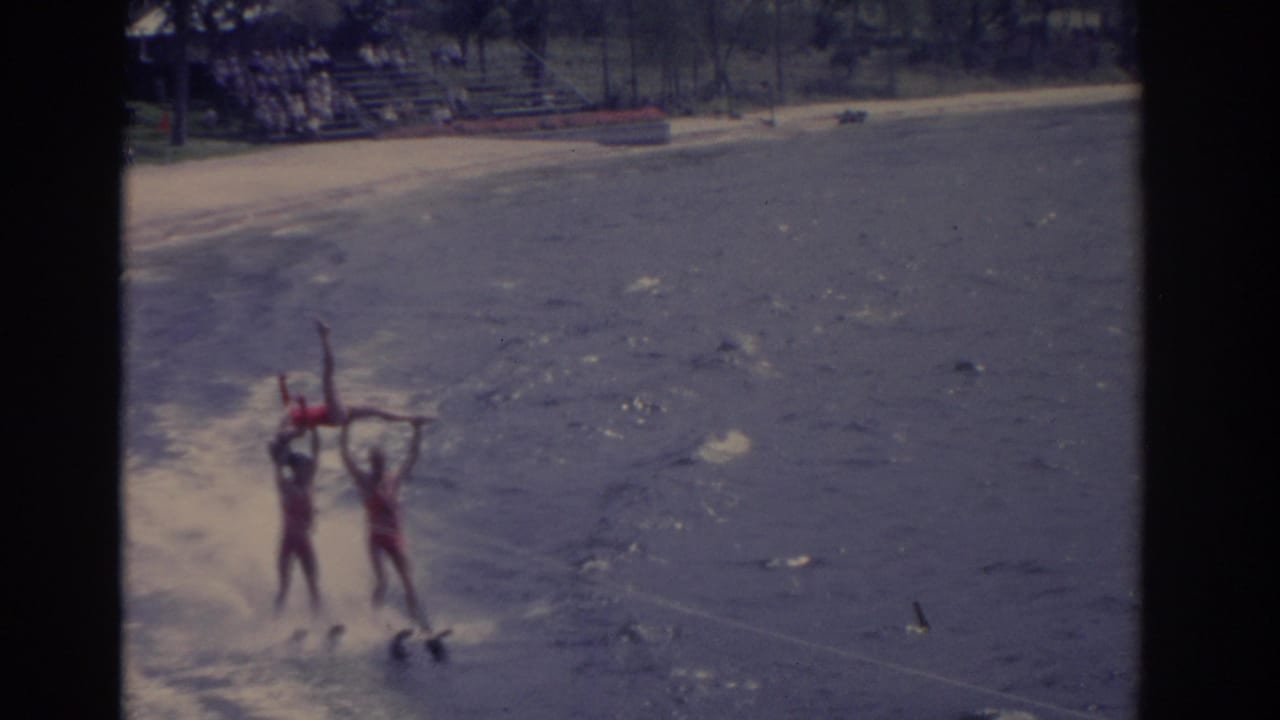 1962 Florida waterski show frame with two red-suited performers lifting another skier above the water as they pass the beach.