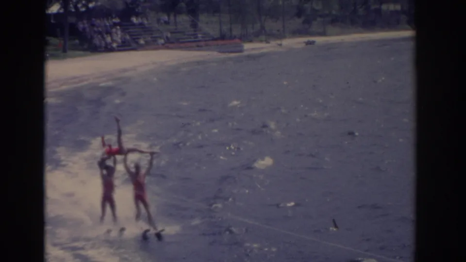 1962 Florida waterski show frame with two red-suited performers lifting another skier above the water as they pass the beach.