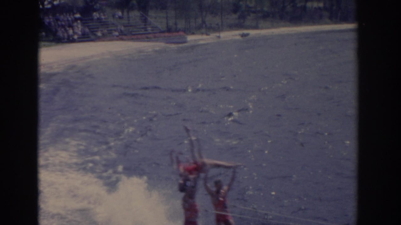 1962 Florida home-movie frame showing a waterski formation low in the frame, with the beach and spectator seating beyond.