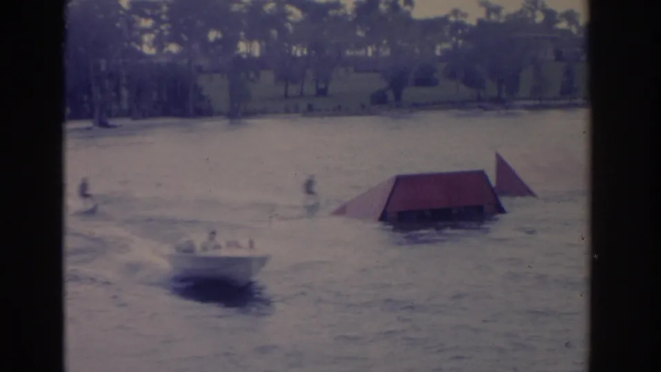 1962 Florida frame showing a boat near a floating ramp, with distant people and trees along the waterline.