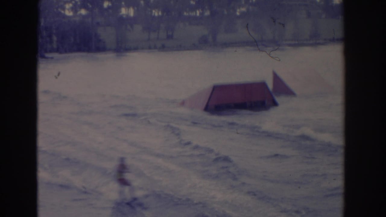 1962 Florida water-ski scene with a distant skier passing near a red ramp on a lake, with trees along the far shore.