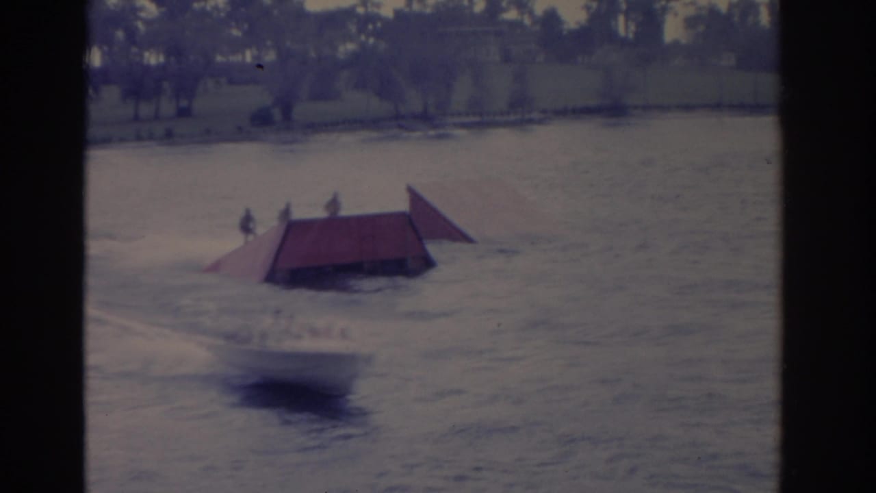 Water skier approaches or passes a red ramp on open water with shoreline in the distance.