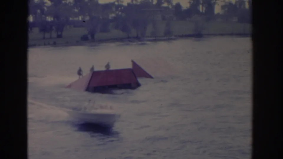 1962 Florida 8mm frame showing a water skier near a red jump ramp on open water, with trees and shoreline beyond.