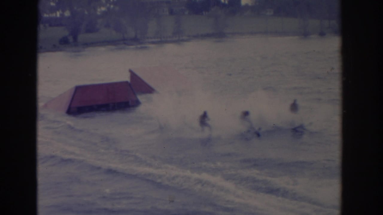 1962 Florida home-movie frame of multiple water skiers passing a red ramp, with spray and wake visible on the water.