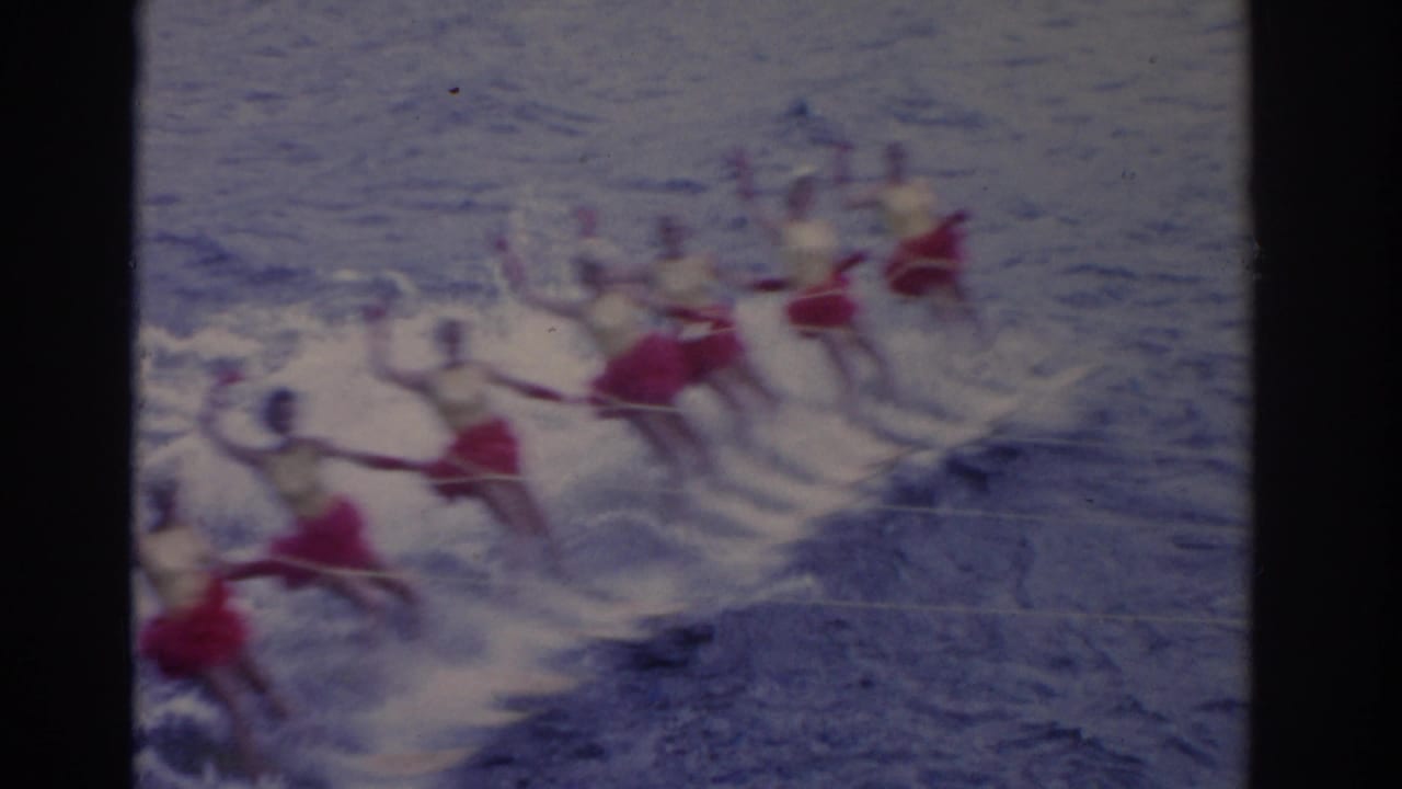 Synchronized water skiers in red skirts skim across a lake in formation.