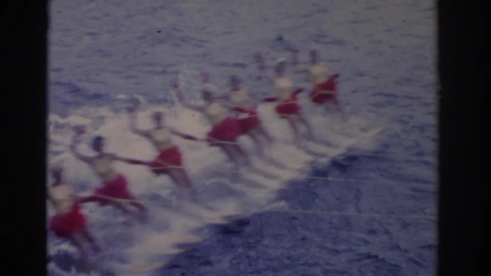 In 1962 Florida footage, a line of water skiers in red skirts and white tops moves across the lake, partly blurred by speed and spray.