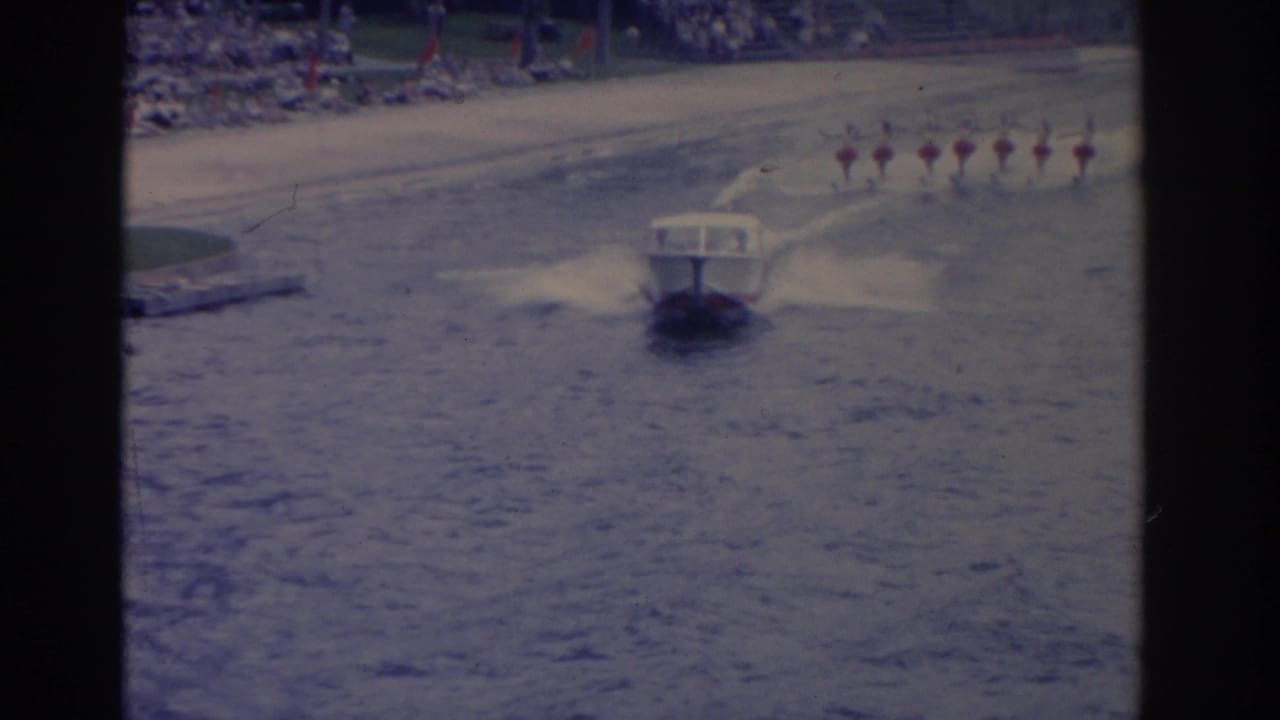 A small motorboat pulls a distant line of synchronized water skiers across a Florida lake, with spectators visible along the shore.