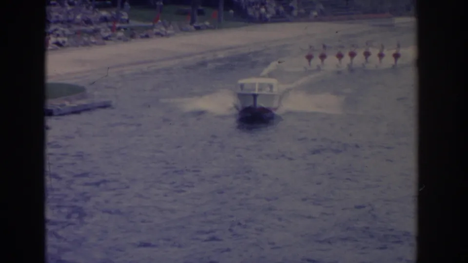 A small motorboat pulls a distant line of synchronized water skiers across a Florida lake, with spectators visible along the shore.