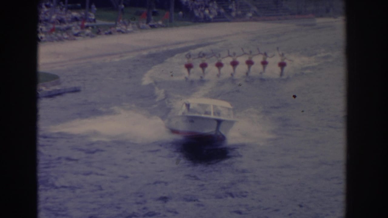 The motorboat speeds through the foreground as the synchronized skiers follow in a line near the beachside audience.