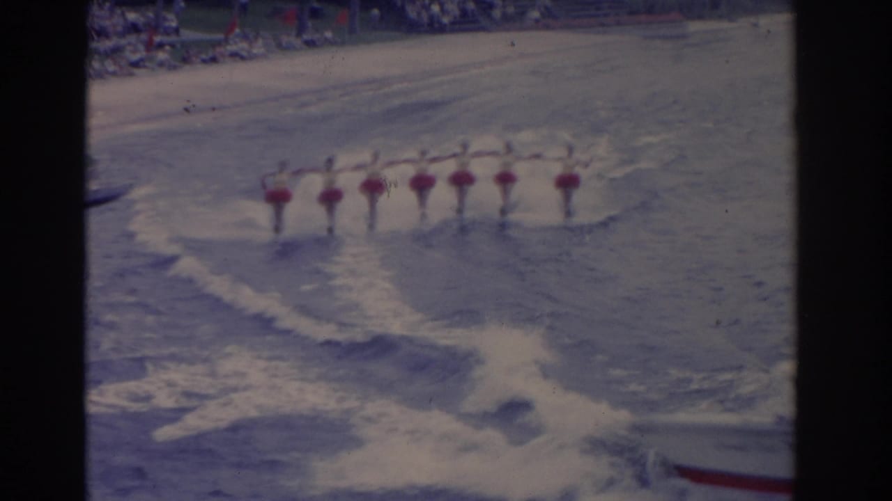 A synchronized line of skiers in red skirts and white tops continues across the lake, framed by wake patterns and faded 8mm color.