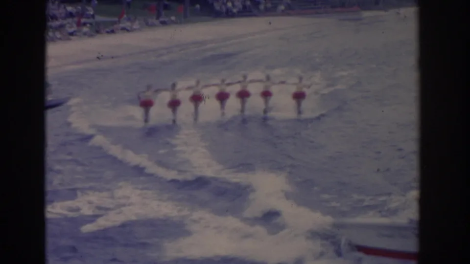 A synchronized line of skiers in red skirts and white tops continues across the lake, framed by wake patterns and faded 8mm color.