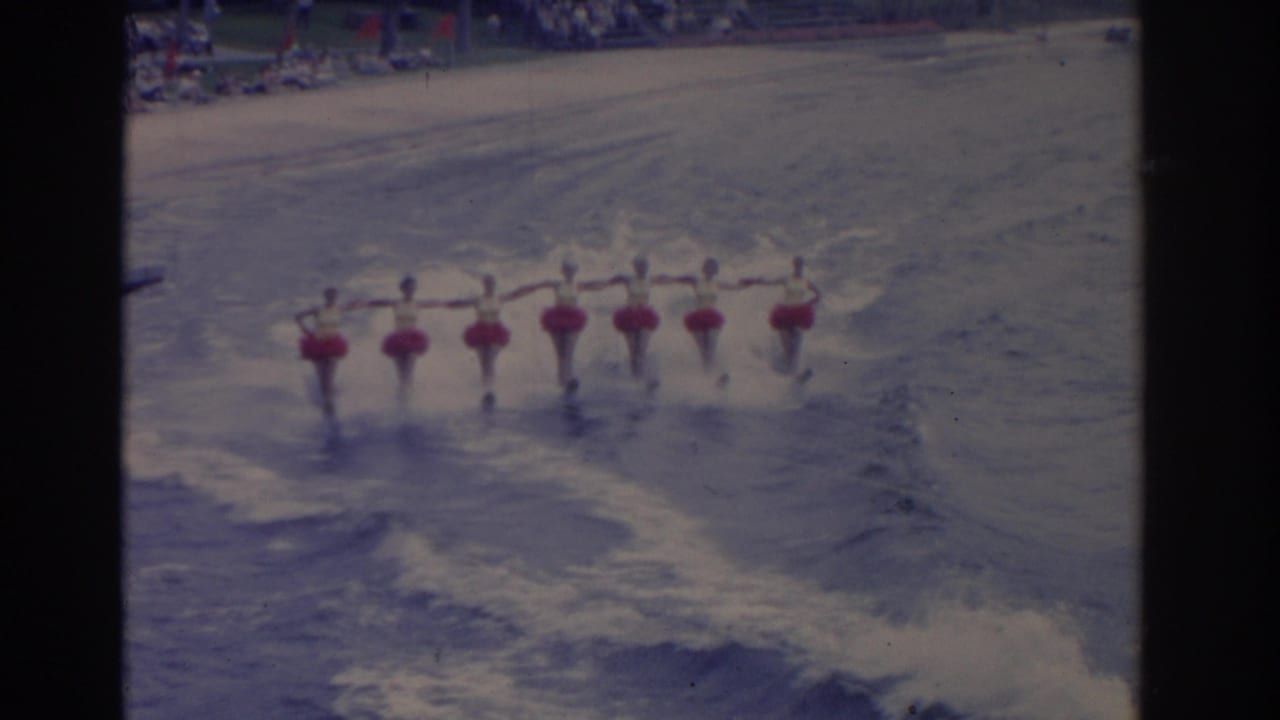 Seven synchronized water skiers hold hands while moving across choppy water.