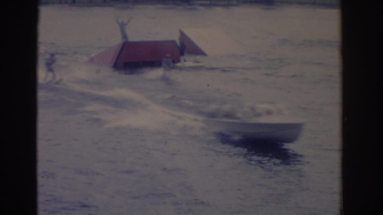 A show platform with red triangular roof panels floats on the water as a performer stands upright above it in the 1962 Florida scene.