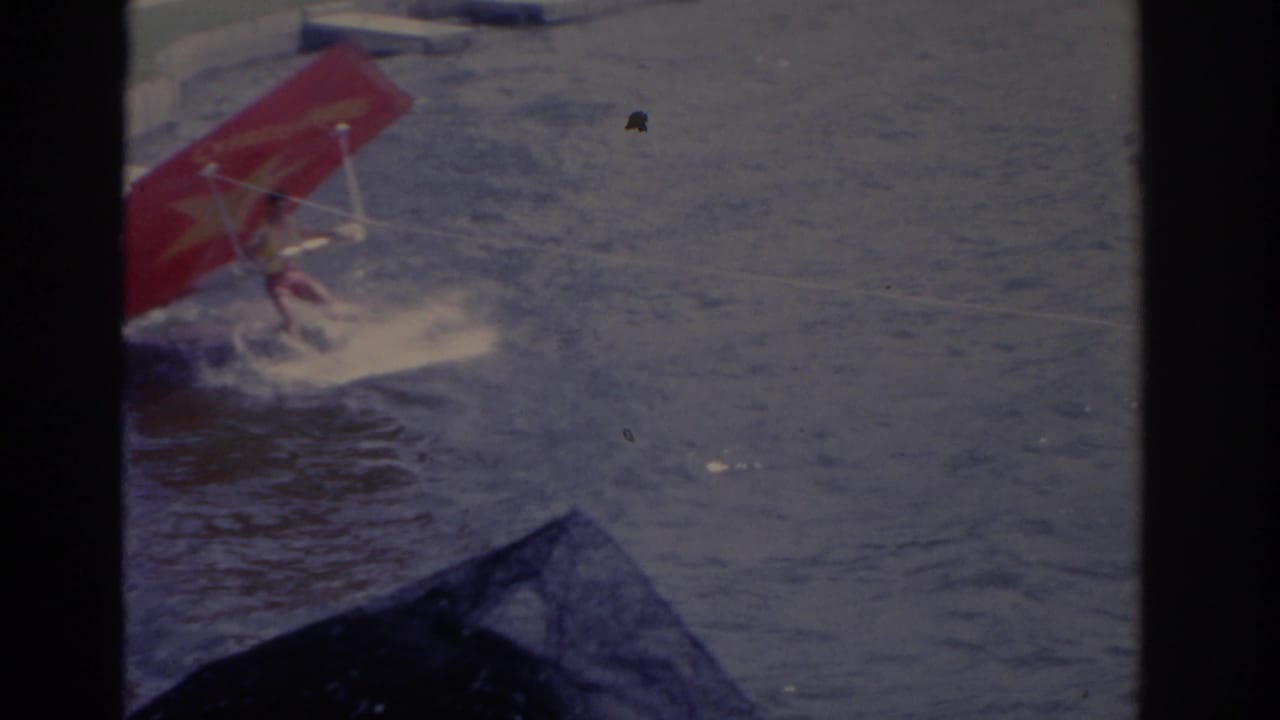 1962 Florida frame of a red sail-like water sport apparatus skimming through choppy water with spray around it.