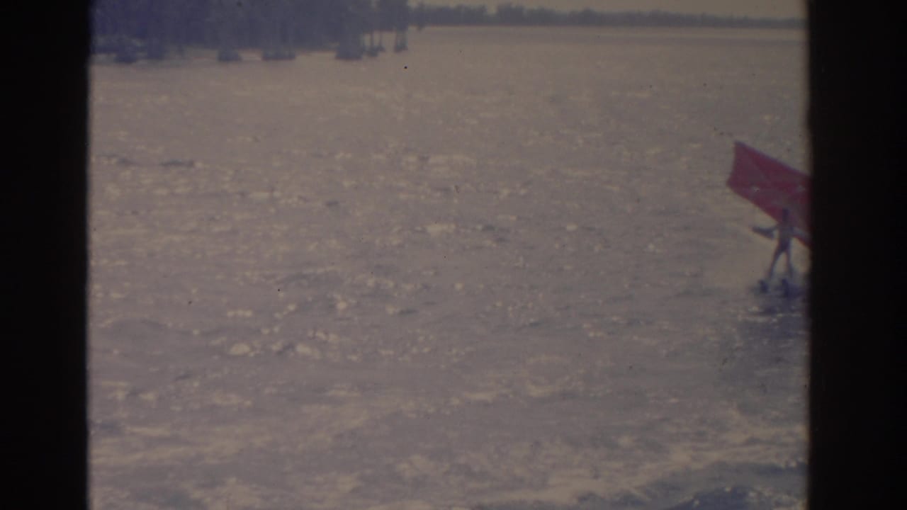 1962 Florida 8mm lake view from a boat, with water, distant trees, a railing, and a small red flag at frame right.