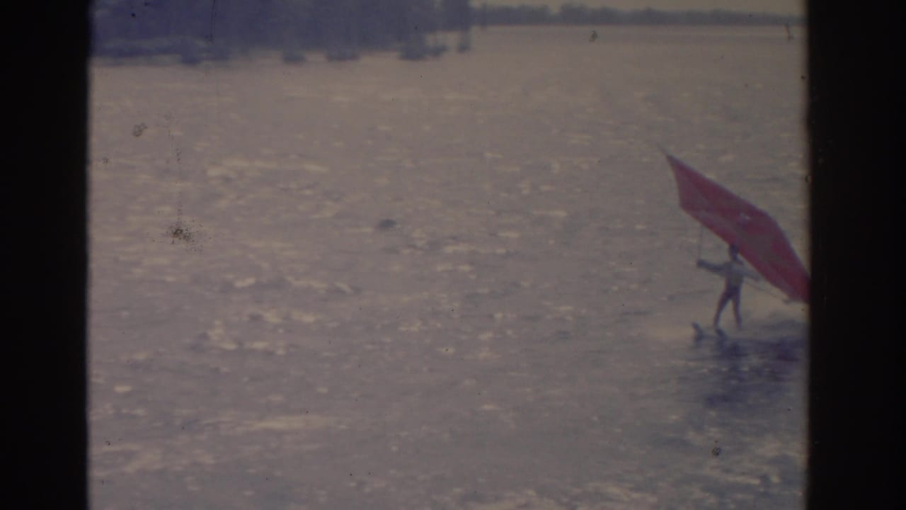 A distant person rides a board with a red sail on a sunlit lake.