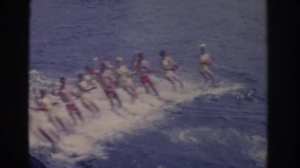A group of people water ski together across a Florida lake, their bright swimwear visible through soft 8mm color and grain.