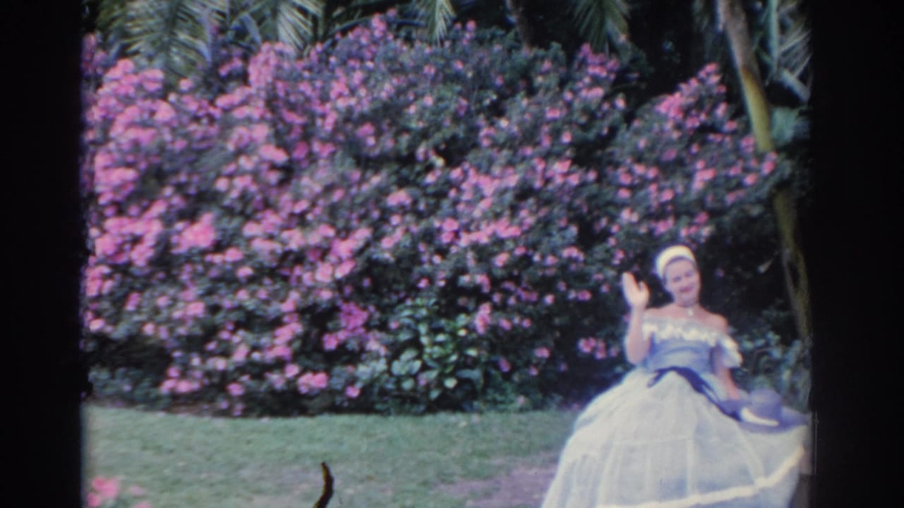 Woman in a white dress waves from a bench in front of pink flowering shrubs.