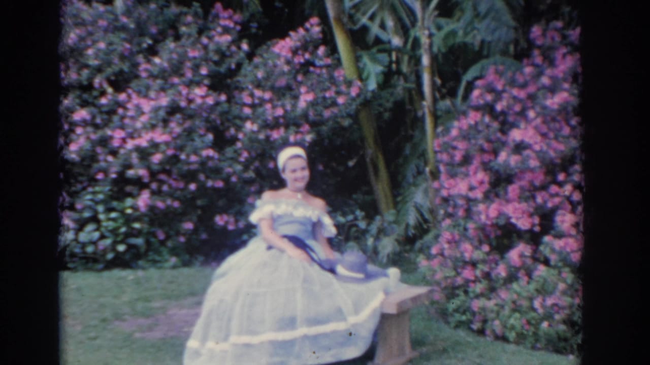 A seated woman in a voluminous white dress is shown near pink flowering bushes and palm-like plants in 1962 Florida footage.