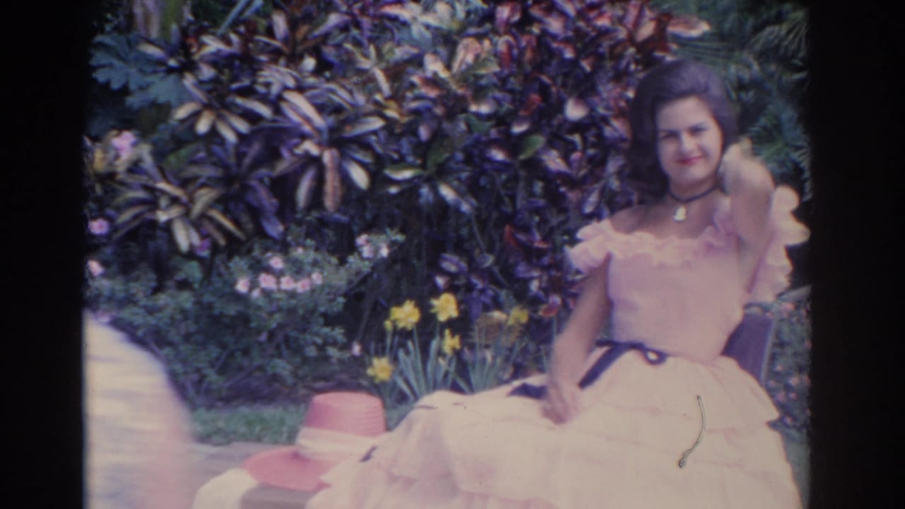 A closer home-movie frame shows the woman smiling in her white dress, with garden foliage, yellow flowers, and a hat nearby.