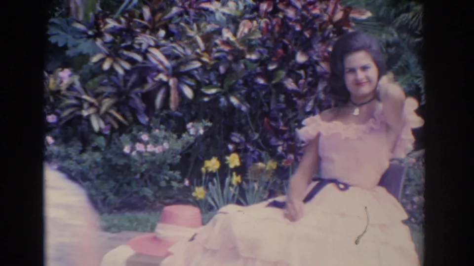A closer home-movie frame shows the woman smiling in her white dress, with garden foliage, yellow flowers, and a hat nearby.