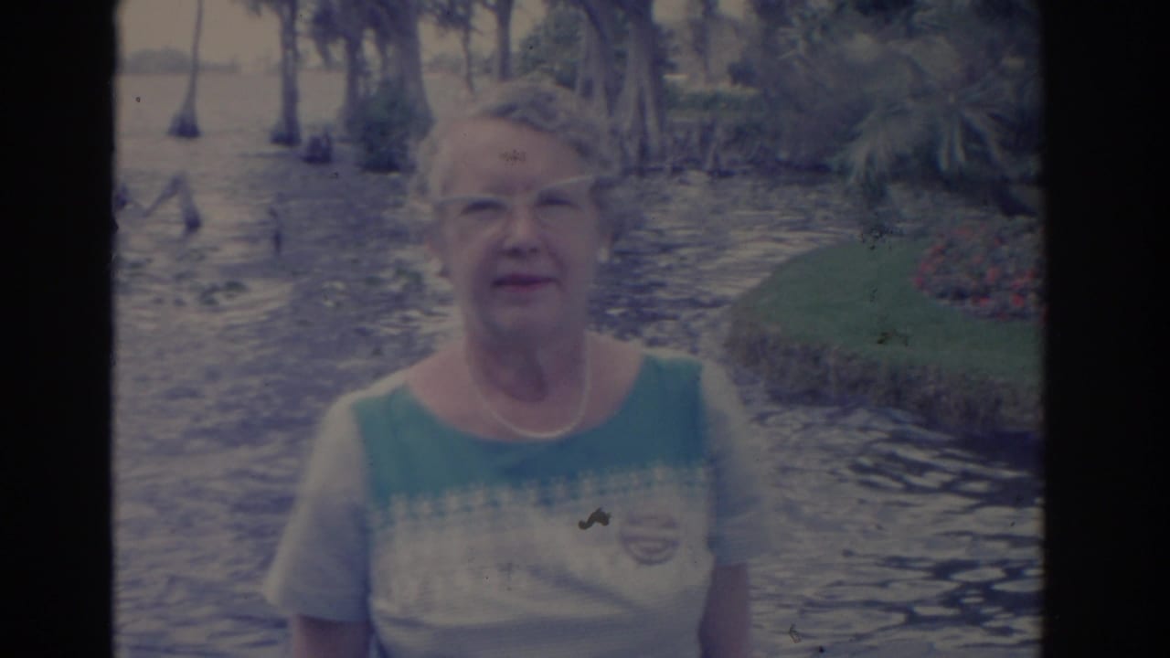 1962 Florida home-movie frame showing an adult woman facing the camera with cypress swamp water and shoreline greenery behind her.